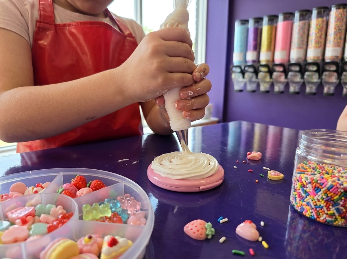 Hands piping white decoden cream onto a pink mirror beside a tray of pastel charms